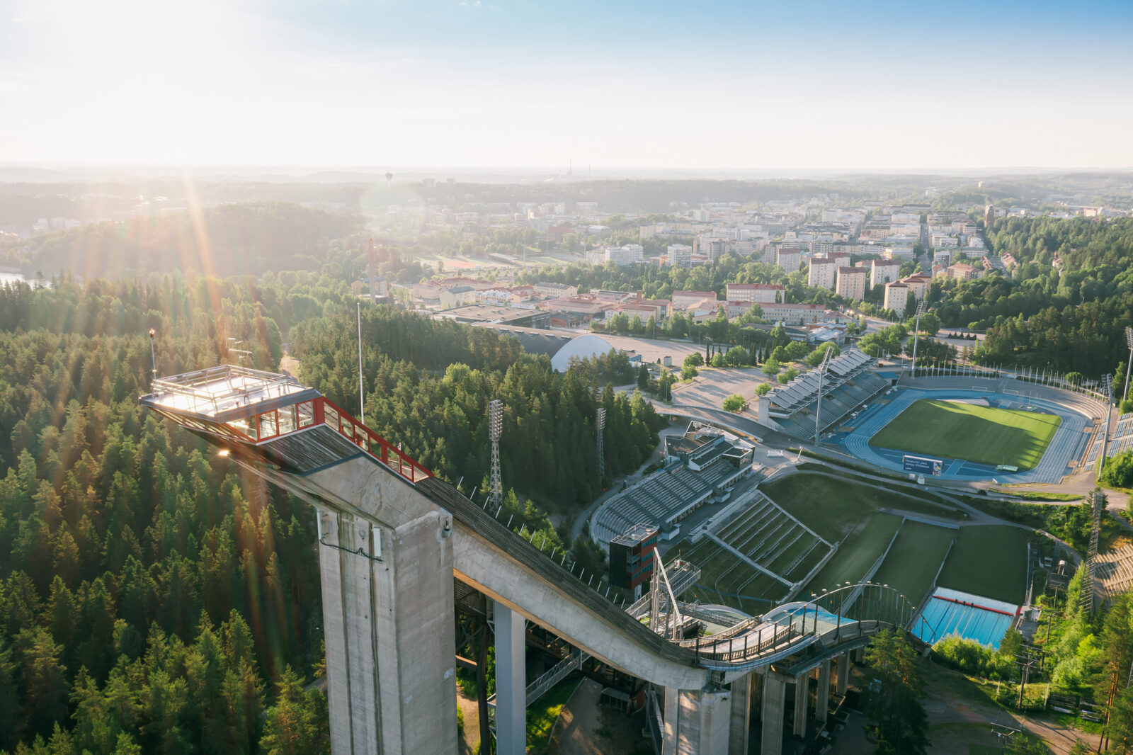 Ilmasta otettu kuva Lahden hyppyrimäestä. Kuvassa näkyy Lahden kaupunkia auringonpaisteessa.
