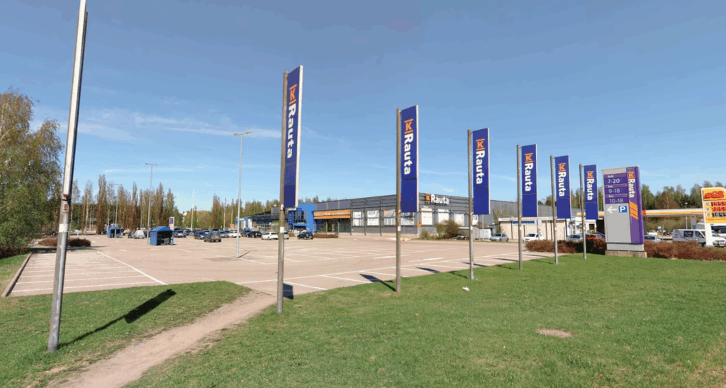 Kuvassa Launeen K-raudan myymälä parkkialueen takana. Taustalla sininen taivas ja etualalla nurmikkoa.